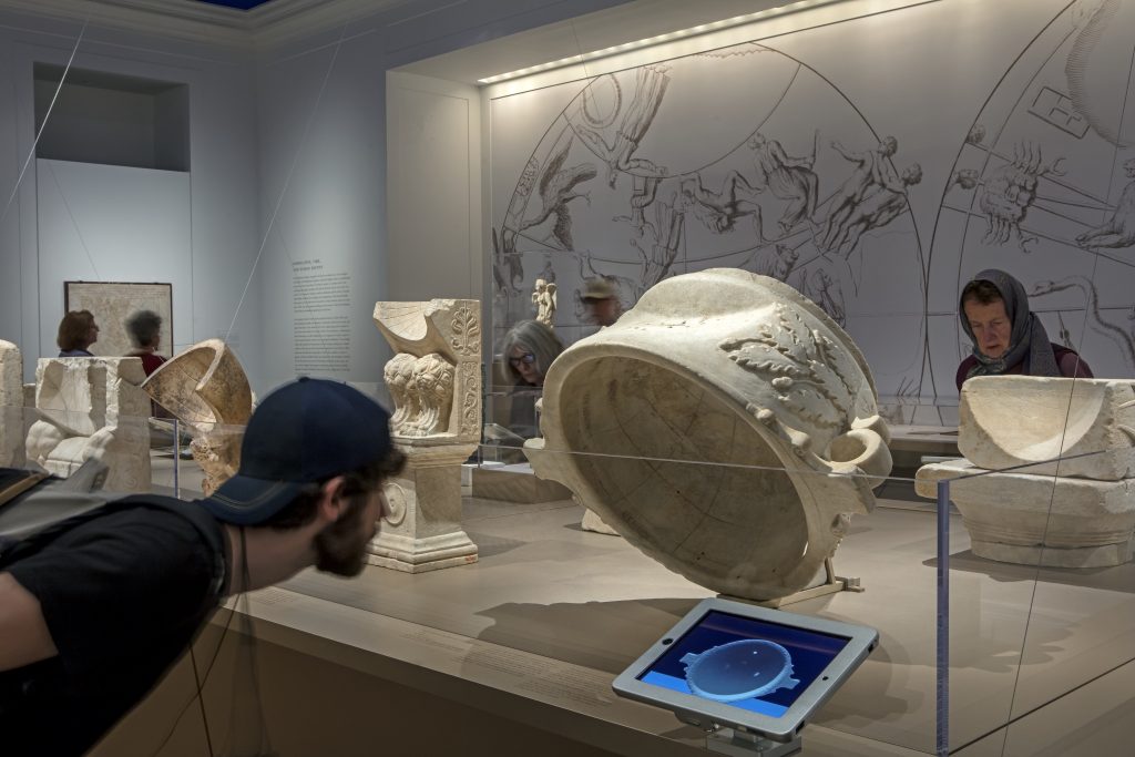 Visitor to the Time and Cosmos exhibition looking closely at the Carthage Roofed Sundial, next to which is an iPad running an animation of shadow on the sundial.