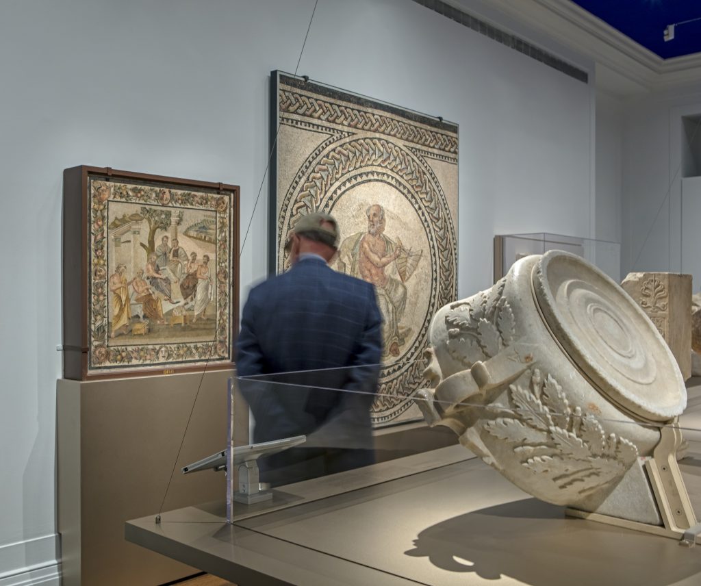 Installation view showing carved exterior of the Roofed Spherical Sundial with Greek Inscription (right) and Roman Mosaic Depicting the Seven Sages (“Plato’s Academy”) on opposite wall (left), 2016. There is a single visitor shown in the image.