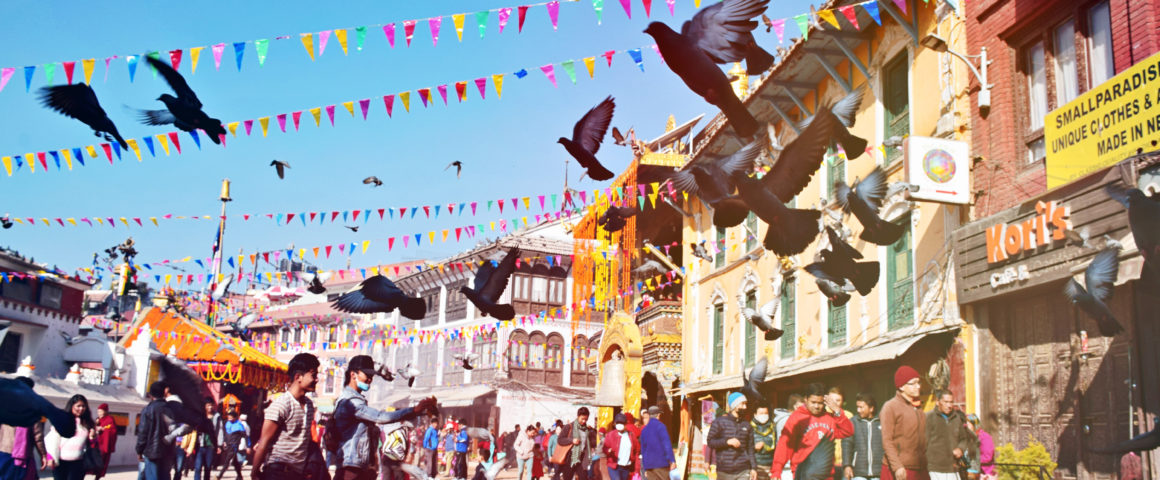 Image depicts the streets of Kathmandu, Nepal. People walk in various directions as birds fly overhead. Pigeons, strollers, and passersby surround Boudhanath Stupa in the morning.