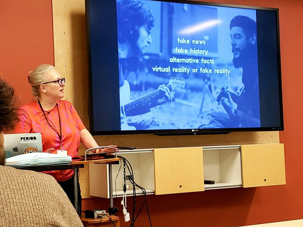 Juliette Levy stands at the podium while presenting a slide reading “fake news, fake history, alternative facts, virtual reality or fake reality” in front of an image of John Lennon and Che Guevara playing the guitar.