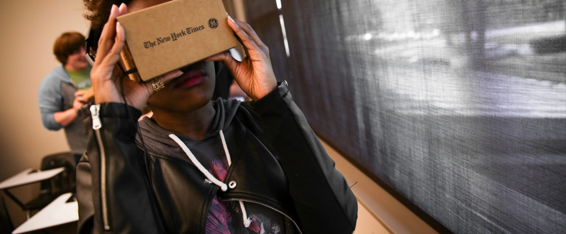 A college student stands by a classroom window wearing a leather jacket holding a Google Cardboard to their face.