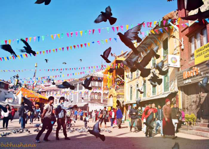 Image depicts the streets of Kathmandu, Nepal. People walk in various directions as birds fly overhead. Pigeons, strollers, and passersby surround Boudhanath Stupa in the morning.