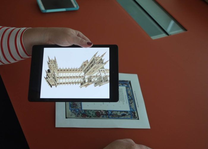 A hand holds a digital tablet over a page of text with a decorative border, while the tablet's screen displays a 3D model of a cathedral.