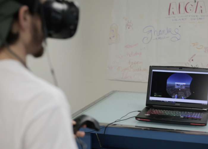 A college student, with their back toward the camera, is wearing a VR headset and holding controllers. In front of them is a gaming laptop with Google Earth displayed. There is a poster with hand drawn words in the background.