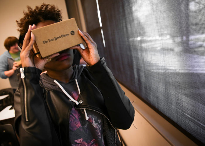 A college student stands by a classroom window wearing a leather jacket holding a Google Cardboard to their face.