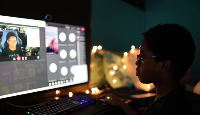 A young person, in shadow and profile, gazes at their own video feed as they take a class online from home, faced with a webcam.