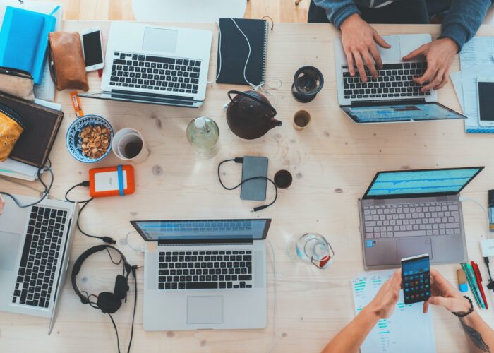 A variety of laptops sit, viewed from above, on a table, worked by hands also using external hard drives, phones, and headphones, alongside snacks and tea.